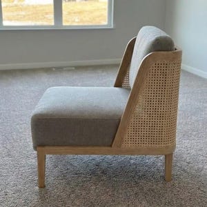 May include: A modern accent chair with a gray upholstered seat and back. The chair features a natural wood frame with woven cane detailing on the back. The chair is set on four wooden legs and is designed for comfort and style.