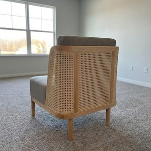 May include: A modern armchair with a light wood frame and woven cane back. The seat and back cushion are upholstered in a gray fabric. The chair has four tapered legs and a curved design, suitable for living room or bedroom decor.