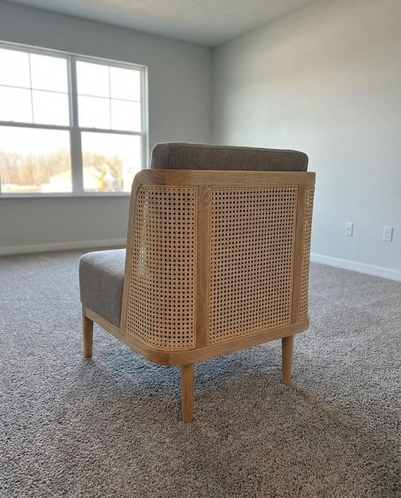 May include: A modern armchair with a light wood frame and woven cane back. The seat and back cushion are upholstered in a gray fabric. The chair has four tapered legs and a curved design, suitable for living room or bedroom decor.