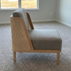 May include: A modern accent chair with a light gray upholstered seat and back. The chair features a natural wood frame and a woven cane back panel. The chair has four wooden legs and is placed on a neutral-colored carpet.