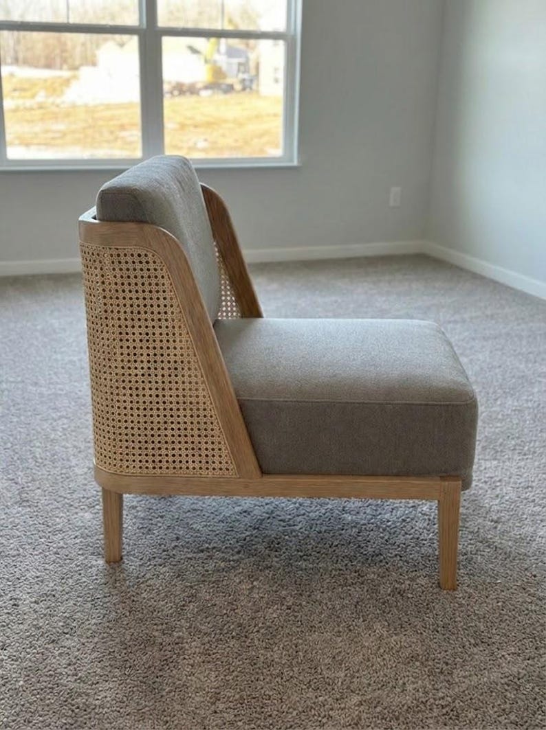 May include: A modern accent chair with a light gray upholstered seat and back. The chair features a natural wood frame and a woven cane back panel. The chair has four wooden legs and is placed on a neutral-colored carpet.