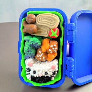 Op de afbeelding: Een blauwe, 3D-geprinte bento-box met een groene binnenkant. De doos is gevuld met miniatuurvoedsel, waaronder een rol, een tomaat, broccoli en een katvormige rijstbal. De doos is open en onthult de inhoud.