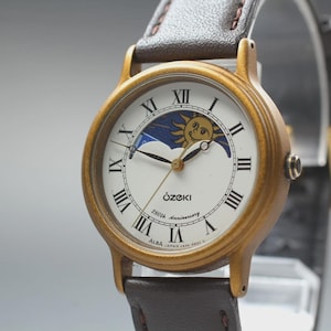 May include: A vintage Ozeki Alba wristwatch with a gold-tone case and a brown leather strap. The white watch face features black Roman numerals, a sun and moon design, and the text "Ozeki 25th Anniversary".