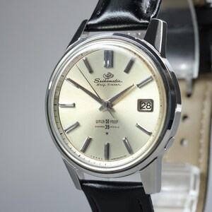May include: A silver-toned Seikomatic Self Dater wristwatch with a champagne-coloured dial, silver hour markers, and a date window at the three o'clock position. The watch features a black leather strap and displays the text Water 50 Proof 39 Jewels.