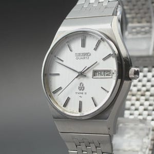 Könnte beinhalten: Eine silberne Seiko Quartz Type II Uhr mit weißem Zifferblatt. Die Uhr hat eine Datumsanzeige mit "DO 2". Sie hat ein silbernes Armband und ein rundes Zifferblatt mit silbernen Stundenmarkierungen.