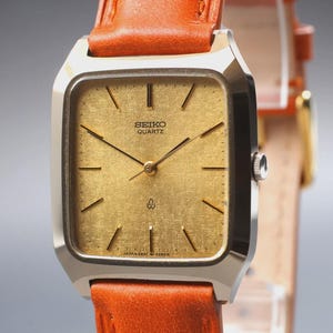 Könnte beinhalten: Vintage Seiko Quartz-Uhr mit rechteckigem, goldfarbenem Zifferblatt und braunem Lederarmband. Die Uhr hat ein silberfarbenes Gehäuse und schwarze Zeiger. Das Zifferblatt zeigt das Seiko Quartz-Logo und Stundenmarkierungen.