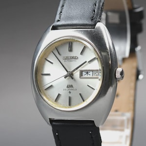 May include: A vintage Seiko automatic wristwatch with a silver dial and black leather band. The watch face displays the day and date, with the text "SEIKO AUTOMATIC" and "23 JEWELS". The watch has a silver-tone case and a black leather strap.