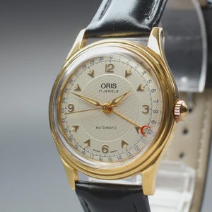 Puede incluir: Un reloj de pulsera Oris en tono dorado con esfera color crema, centro texturizado, números arábigos en 3, 6, 9 y 12, aguja de fecha con punta de media luna roja y correa de cuero negro con hebilla pulida en tono dorado.