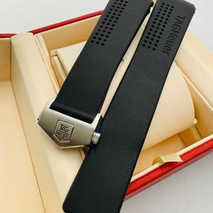 May include: Black watch strap with the text "TAG Heuer" and a silver buckle. The strap has a perforated design and is displayed inside a red and beige box.