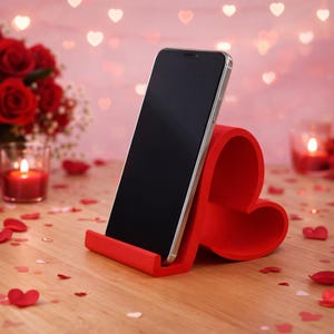 May include: A red heart-shaped phone stand holding a smartphone. The stand is made of a solid material and has a slot to hold the phone upright. The background includes red rose bouquets, candles, and heart-shaped confetti.