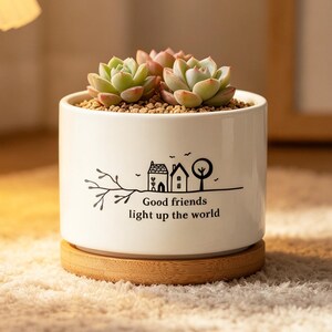 May include: A white ceramic plant pot with a succulent, on a wooden coaster. The pot features a black illustration of houses, trees, and the text "Good friends light up the world".