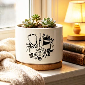 May include: A white ceramic planter with a black graphic of medical tools, coffee, and flowers, with the name "Ashley" on a banner. The planter holds several green succulents and sits on a wooden base. A small lamp and books are in the background.