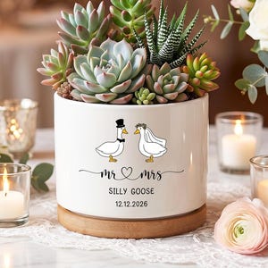 Op de afbeelding: Een witte keramische plantenbak met vetplanten, met een Mr. en Mrs. ganzenontwerp. De plantenbak staat op een houten voet en bevat de tekst "Mr & Mrs", "Silly Goose" en de datum 12.12.2026.