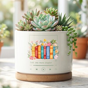 May include: A white ceramic planter with a wooden base, filled with various succulents. The planter features a colorful illustration of books and flowers, with the text "Just one more chapter".