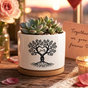 Può includere: Un vaso in ceramica bianca per piante grasse con base in bambù, caratterizzato da una grafica nera di un albero con tronco a forma di cuore contenente le lettere J+S e la data 10.10.2026, contenente una pianta grassa verde.