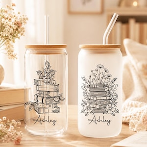 Könnte beinhalten: Zwei Trinkgläser mit Bambusdeckeln und klaren Strohhalmen. Ein Becher ist klar, der andere mattiert. Beide zeigen einen Stapel Bücher mit floralen Akzenten und dem Namen "Ashley". Ideal für Eiskaffee oder Tee.