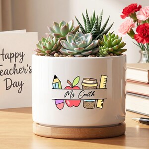 May include: White ceramic planter with a teacher-themed design, holding several succulents. The planter features a pencil, apple, coffee cup, and ruler graphic, with the name "Ms. Smith" below. A wooden base supports the planter. A card reads "Happy Teacher's Day".