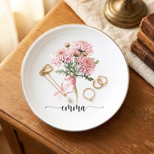 Op de afbeelding: Wit keramisch sieradenbord met een gouden rand, met een boeket roze bloemen en de naam "emma". Een gouden ketting en ringen liggen op het bord, perfect voor het opbergen van kleine accessoires.