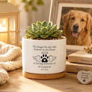 Könnte beinhalten: Ein weißer Keramikübertopf mit einer grünen Sukkulente, bedruckt mit schwarzem Text: No longer by my side forever in my heart, in loving memory of Cooper, 2014-2026, sowie einer Pfotenabdruck- und Engelsflügelgrafik, auf einem runden Holzsockel.
