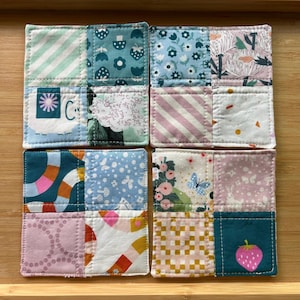 May include: Set of four square fabric coasters with a patchwork design. Each coaster features a unique combination of patterned fabrics in shades of blue, pink, green, and white. Designs include stripes, florals, and geometric shapes. The coasters are likely handmade.