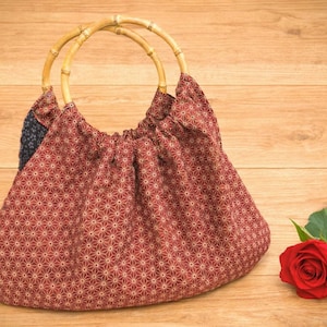 以下が含まれることがあります： 竹製の持ち手が付いた赤い模様のハンドバッグが、赤いバラの隣の木製の表面に置かれています。バッグは、繰り返しのスターバーストデザインが特徴です。ハンドルは、湾曲した明るい色の竹で作られています。