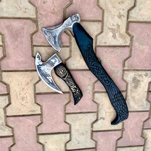 May include: Two Viking axes with intricate designs. The larger axe has a silver blade and a dark blue handle with Celtic knotwork. The smaller axe has a silver blade with runes and a black handle with spiral and line patterns.