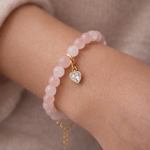 May include: Rose quartz bracelet featuring round, light pink beads. A gold-colored charm with a heart-shaped, clear crystal pendant hangs from the bracelet. The text "ROSE QUARTZ BRACELET" is at the top of the image.