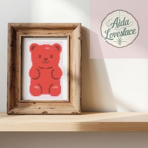 May include: A framed cross-stitch of a red gummy bear. The artwork is displayed in a rustic wooden frame and set against a light pink background. The frame sits on a light-coloured wooden shelf. The logo "Aida Lovelace" is visible in the upper right corner.