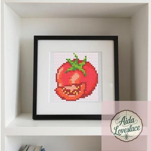 May include: Cross-stitch art of a red tomato with green leaves, framed in black. The tomato is detailed with various shades of red and orange, showcasing a textured appearance. The artwork is displayed on a white shelf.