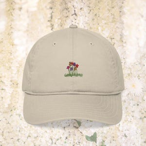 May include: A beige baseball cap with a curved peak. The cap has an embroidered design of colourful flowers and green grass. The hat is made of a soft material and has a classic, casual style.