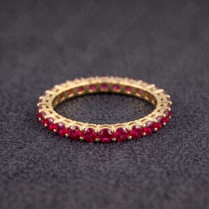Op de afbeelding: Een gouden eeuwigheidsring met een doorlopende rij ronde, levendig rode robijnen in een gedeelde klauwzetting. De ring heeft een cirkelvormig ontwerp met een uniforme plaatsing van de edelstenen en gepolijst goudkleurig metaal.