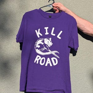 Könnte beinhalten: Lilafarbenes T-Shirt mit dem weißen Schriftzug "KILL ROAD". Zwischen den Wörtern befindet sich eine weiß-graue Opossum-Grafik. Das T-Shirt hängt an einem schwarzen Kleiderbügel.