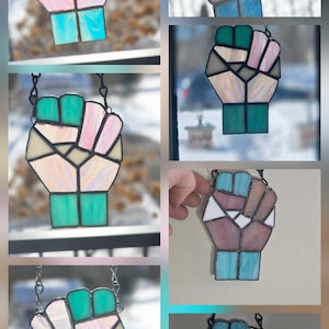 May include: A stained glass fist pendant in various colors, including pink, blue, green, and white. The pendant is made of multiple geometric shapes and hangs from a silver chain. The fist is a symbol of solidarity and resistance.