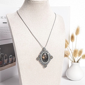 May include: A silver-toned pendant necklace with an oval-shaped, dark-colored stone set within a decorative, macrame-style frame. The pendant hangs from a delicate chain. The necklace is displayed on a mannequin bust.