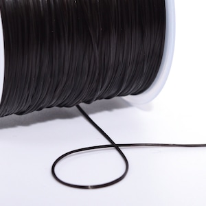 Puede incluir: Una bobina de cordón elástico negro, enrollada apretadamente en una bobina de plástico blanco. Una longitud del cordón se extiende desde la bobina, formando un bucle.