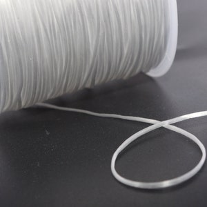 Pode incluir: Uma bobina de cordão elástico transparente, enrolada em torno de uma bobina de plástico branco. O cordão está sobre uma superfície escura.