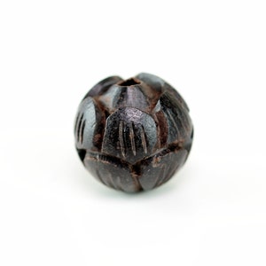 May include: A dark brown wooden bead with a carved, geometric pattern. The bead is round and has a small hole in the center.