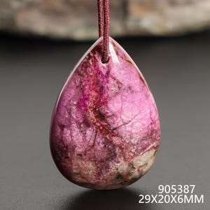 May include: A teardrop-shaped pendant with a polished, marbled appearance. The stone features shades of pink, red, and brown with a visible grain. The pendant is suspended by a maroon cord. The dimensions are 29x20x6MM.