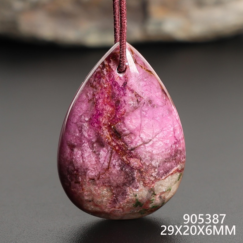 May include: A teardrop-shaped pendant with a polished, marbled appearance. The stone features shades of pink, red, and brown with a visible grain. The pendant is suspended by a maroon cord. The dimensions are 29x20x6MM.