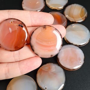 May include: A collection of polished, round agate stones in shades of orange, brown, and white. Each stone has a hole drilled through it. The stones are displayed on a dark surface.