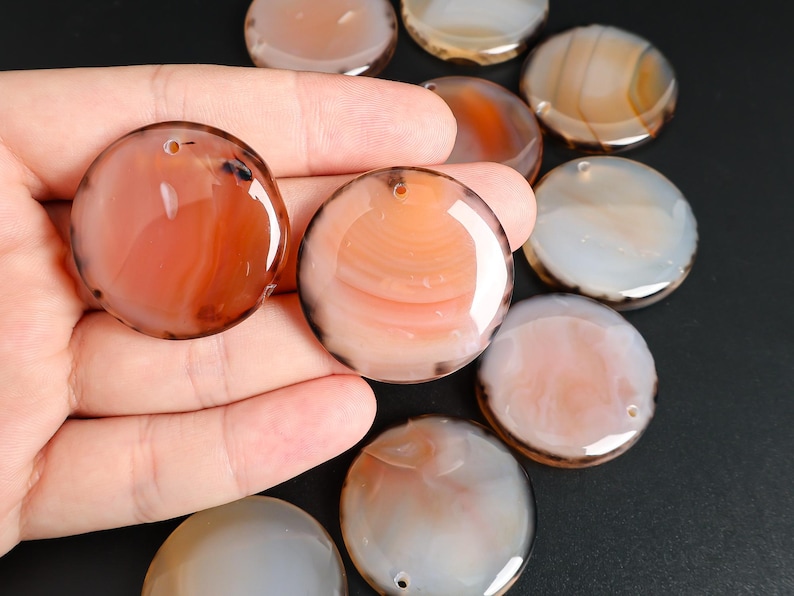 May include: A collection of polished, round agate stones in shades of orange, brown, and white. Each stone has a hole drilled through it. The stones are displayed on a dark surface.