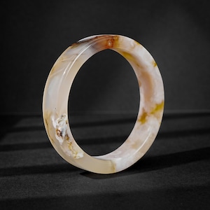 May include: A polished bangle bracelet crafted from translucent agate stone. The circular band features a smooth, glossy surface with natural swirling patterns in shades of cream, beige, soft brown, and hints of golden yellow throughout the stone.