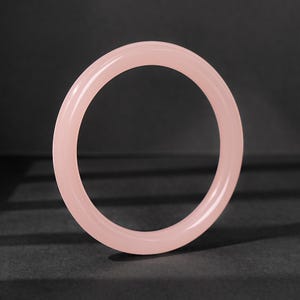 May include: A smooth, solid light pink bangle bracelet with a polished, translucent finish. The circular jewelry piece features a rounded profile and a consistent thickness throughout its entire circumference, showcasing a simple and clean design.