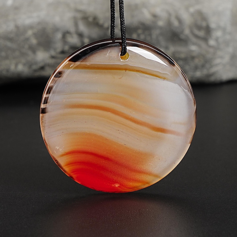 May include: A round pendant with a natural agate design. The pendant features layers of white, orange, and red hues, creating a marbled effect. It is suspended from a black cord, suitable for wearing as a necklace. The stone is polished and has a glossy finish.