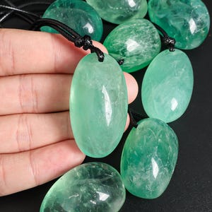May include: A collection of oval-shaped, translucent green fluorite pendants on black cords. The pendants are held in a hand, showcasing their polished surfaces and natural variations in color and clarity. The background is a dark, solid color.