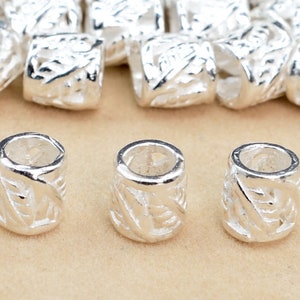 May include: Silver metal beads with a decorative design. The beads are cylindrical and have a hollow center.
