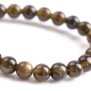 Puede incluir: Primer plano de una pulsera de cuentas redondas pulidas en tonos marr&oacute;n y dorado. Las cuentas tienen un brillo met&aacute;lico reflectante, lo que sugiere un material de piedra preciosa natural. La pulsera est&aacute; ensartada en un cord&oacute;n flexible. Di&aacute;metro aprox. 1 cm.