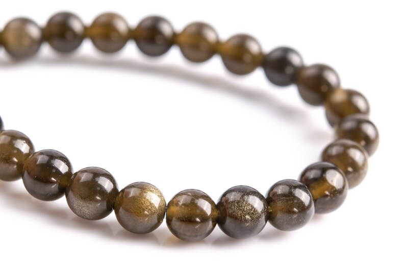 Puede incluir: Primer plano de una pulsera de cuentas redondas pulidas en tonos marr&oacute;n y dorado. Las cuentas tienen un brillo met&aacute;lico reflectante, lo que sugiere un material de piedra preciosa natural. La pulsera est&aacute; ensartada en un cord&oacute;n flexible. Di&aacute;metro aprox. 1 cm.