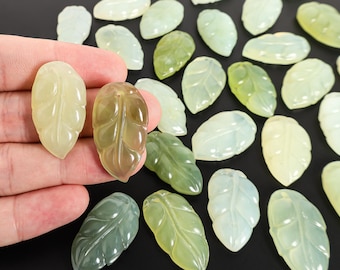 May include: A collection of leaf-shaped gemstone pendants in various shades of green and yellow. Two pendants are held in a hand, showcasing their detailed veining. The other pendants are scattered, creating a natural, organic aesthetic. These pendants are ideal for jewelry making.