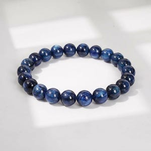 Puede incluir: Una pulsera elástica compuesta por cuentas redondas y pulidas de cianita. Las cuentas presentan un color azul profundo con sutiles vetas azul claro y un acabado brillante y reflectante, dispuestas en un bucle circular continuo.